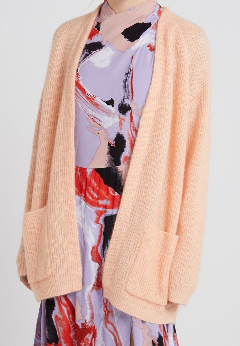 Peach fuzzy cardigan with long sleeves, open front, and two front pockets, layered over a multicolored abstract print dress.