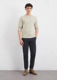 Chemise beige à manches longues avec un col rond côtelé, associée à un pantalon chino marine et des bottines en suède tan à lacets, se tenant sur un fond neutre.
