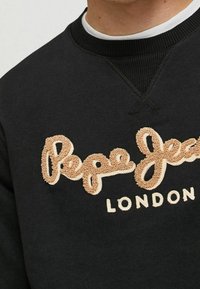 La sudadera negra presenta un texto bordado "Pepe Jeans London" en beige y marrón claro, con un cuello de canalé y un acabado texturizado.