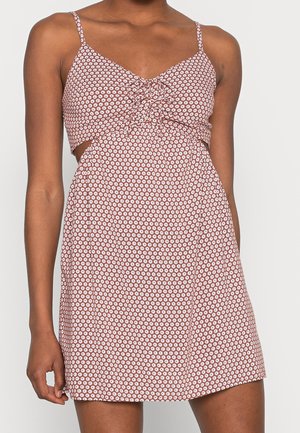 Robe marron sans manches avec imprimé floral blanc, devant froncé et découpes latérales portée par une personne à la peau foncée.