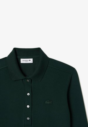 Polo de color verde oscuro con una tela texturizada, un cuello y tres botones. Presenta un logo bordado de Lacoste en el lado izquierdo.