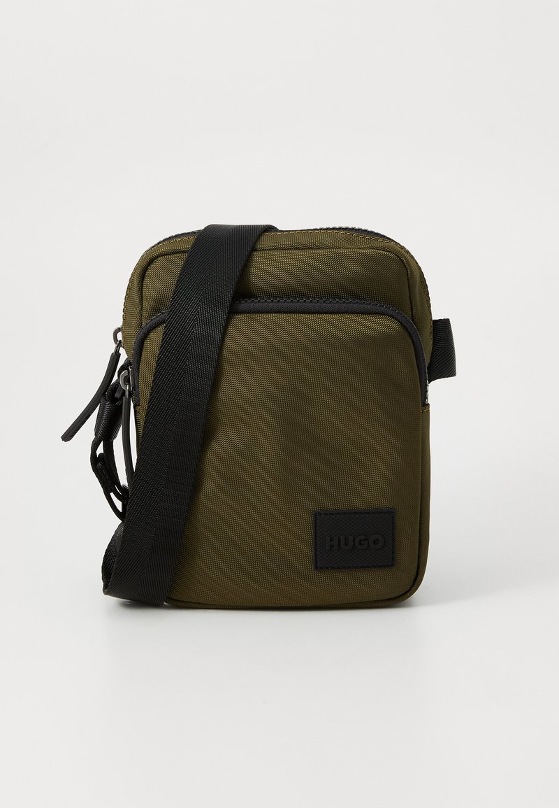 Olivengrønn nylon crossover-veske med en svart stropp, med glidelås og en frontlogo-patch. Teksturert overflate og rektangulær form.