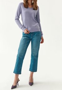 Sweter w kolorze lawendy z dekoltem w serek, o miękkiej dzianinie. Noszony z jasnoniebieskimi jeansami o prostych nogawkach i fioletowymi szpilkami. Prosty design i fason.