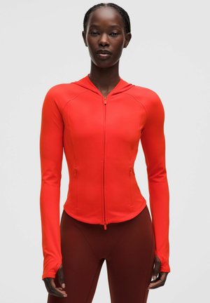 Femme portant un sweat à capuche rouge vif ajusté avec fermeture éclair, trous pour les pouces et leggings rouge foncé, debout devant un fond clair uni.