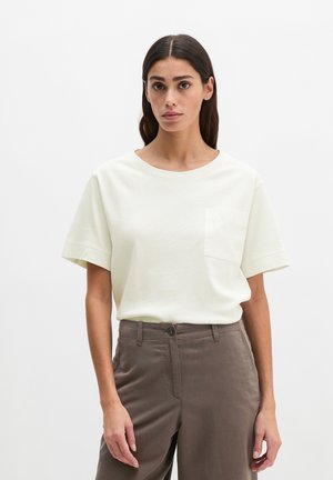 Vrouw staande tegen een witte achtergrond, gekleed in een crèmekleurig shirt met korte mouwen en een zak, en een bruin, hooggetailleerd broek, voorwaarts gericht.