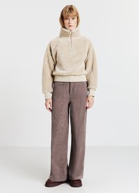 Maglione beige in pile con collo alto e zip a quartino; abbinato a pantaloni marroni in velluto a coste larghi e testurizzati. Scarpe basse con suole morbide.