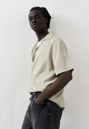 Jeune homme à la peau foncée et aux dreadlocks portant une chemise beige à manches courtes boutonnée et un jean noir, debout contre un mur blanc.