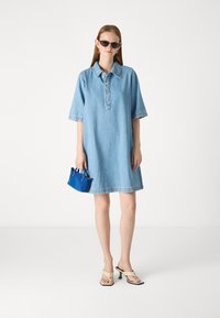 Denim dress in light blue with short sleeves, buttoned collar, and raw hem. Accompanied by a small blue handbag and beige heeled sandals.