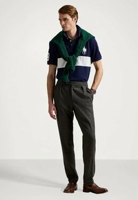 Granatowa koszulka polo z białymi paskami, zielony sweter dzianinowy na ramionach, szare teksturowane spodnie i brązowe skórzane buty. Swobodny strój z klasycznymi detalami.