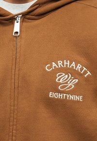 Hnedá mikina na zips s bielym vyšívaným textom "Carhartt Wip Eightynine" na hrudi, nosená cez biele tričko.