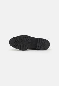 Semelle de chaussure noire en caoutchouc avec des profondeurs de crampons et un logo en relief, présentant une surface texturée pour l'adhérence et la stabilité.