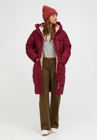 Bordeauxkleurige pufferjas met roze voering, grote capuchon en drukknoopsluiting. Gecombineerd met een grijs shirt en olijfgroene broek, gedragen met witte schoenen.