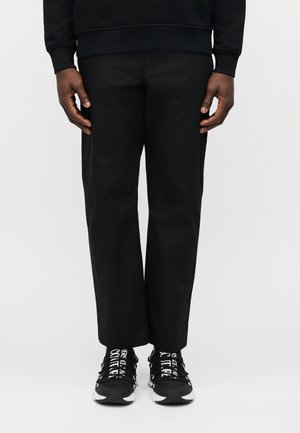 Oseba, oblečena v črne hlače s ravnimi hlačnicami in črne superge z belim napisom na vezalkah, stoji ob enobarvnem belem ozadju.