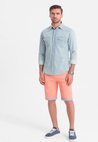 Camicetta in denim azzurro chiaro con due tasche sul petto, abbinata a pantaloni corti corallo chiaro con risvolti. Indossata con sneakers blu.