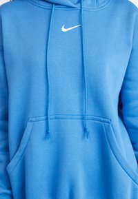 Blå hoodie av mjukt tyg med en känguruficka och dragsnören, med en vit Nike-logotyp på bröstet. Horisontell textur synlig.