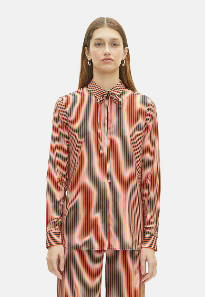 Vrouw in overeenkomstige gestreepte shirt met lange mouwen en broek in rood, zwart en beige met een strik-kraag, staande tegen een witte achtergrond.