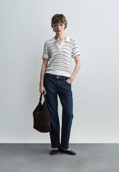 Γυναίκα που φοράει πλεκτό πόλο μπλουζάκι με κοντά μανίκια σε λευκό χρώμα, σκούρο μπλε τζιν, μαύρες μπαλαρίνες, κρατώντας μεγάλη σκούρα καφέ τσάντα, στέκεται μπροστά σε απλό φόντο.