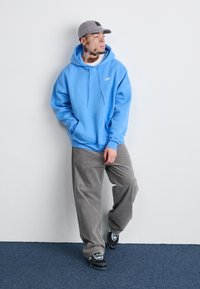 Blaues Kapuzensweatshirt mit weißem Logo, graue Cordhosen und gemusterte schwarze Sneakers. Stehend vor einem einfarbigen Hintergrund.