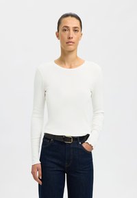 Top de manga larga blanco con textura acanalada y cuello redondo, combinado con jeans de mezclilla azul oscuro y un cinturón negro con hebilla dorada.