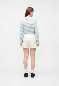 Γυναίκα που έχει την πλάτη γυρισμένη, φοράει μπλε γαλάζιο και λευκό ριγέ μακρυμάνικο πουκάμισο, κρεμ σορτς, λευκές κάλτσες και μαύρα αθλητικά παπούτσια σε λευκό φόντο.