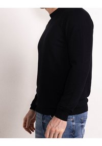 Uomo che indossa un maglione nero a maniche lunghe e jeans blu, in piedi di lato contro un muro bianco uniforme.