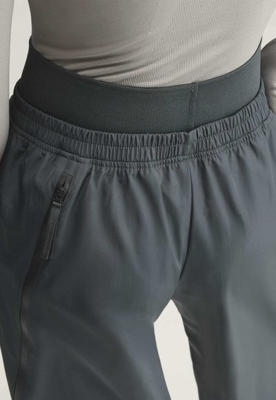 Nahaufnahme von dunkelgrauen Hosen mit elastischem Bund und seitlicher Reißverschlusstasche, kombiniert mit einem hellgrauen, taillierten Oberteil.