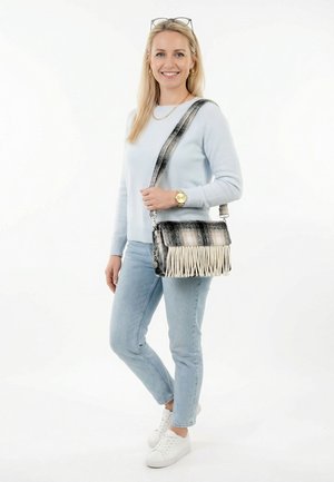 Sieviete gaiši zilā džemperī un džinsos, nēsājot melnas un baltas bārkstis plecu somu, valkājot baltas sporta kurpes un zelta pulksteni.