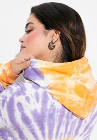 Hoodie tie-dye con colori arancione, viola e bianco. Tessuto morbido con cappuccio regolabile e vestibilità comoda, indossato di lato.