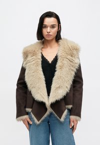 Καφέ σουέντ μπουφάν με μπεζ τεχνητή γούνα στο γιακά και τα περιμετρικά μέρη. Διαθέτει διαγώνιο τελείωμα και λεπτομέρειες με κουμπιά μπροστά. Φορεμένο πάνω από μαύρο τοπ.