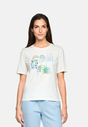 Frau trägt ein weißes T-Shirt mit abstrakten blauen und grünen Streifen sowie hellblaue, strukturierte Hosen mit Seitenstreifen, steht vor weißem Hintergrund.