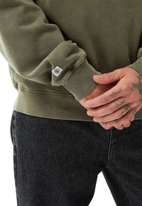 Person med tatuerade händer i en olivgrön sweatshirt och svarta jeans, som visar sweatshirtens ärm med en liten logotypetikett.