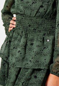 Zielona sukienka z lekkiego materiału, z marszczonym pasem, z czarnym wzorem gwiazd i planet oraz małym metalicznym akcentem.