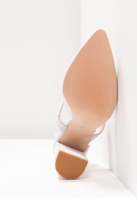 Vit högklackad sko med spetsig tå, med en texturerad mockaöverdel och en ljusbrun gummisula med vertikala spår.