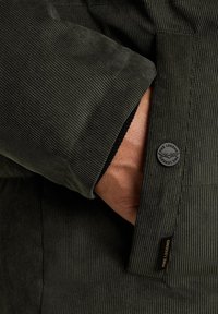 Donkergroene corduroy jas met geribbelde textuur. Heeft een strakke manchet en een zwarte merkbutton bij de zak voor extra detail.