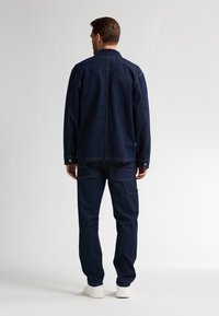 Uomo in piedi con la schiena rivolta, indossa una giacca di jeans blu scuro e pantaloni con tasche, e sneakers bianche su uno sfondo semplice.