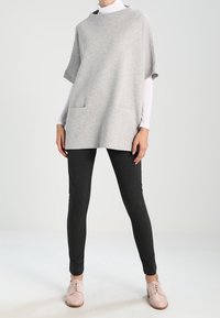 Lichtgrijze poncho-stijl top met ribbeltextuur en twee voorkwanten, gecombineerd met zwarte taps toelopende broek en lichtroze veterschoenen.