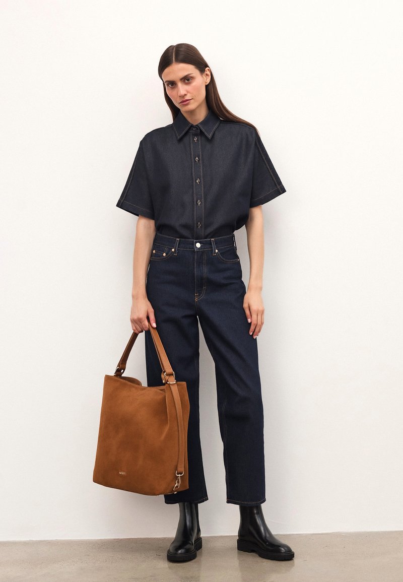 Dunkelblaues Denim-Hemd mit kurzen Ärmeln und Knopfdetails, kombiniert mit hochgeschnittenen dunklen Jeans. Braune Wildleder-Tasche mit Schultergurt. Schwarze Stiefeletten.