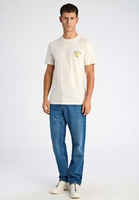 T-shirt crema a maniche corte con una grafica gialla sul petto, abbinata a jeans blu a gamba dritta e sneakers bianche.