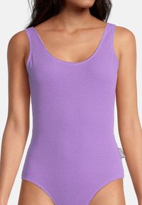 Femme portant un maillot de bain sans manches texturé lilas avec un décolleté bateau et une petite étiquette sur le côté portant la mention "BECO".