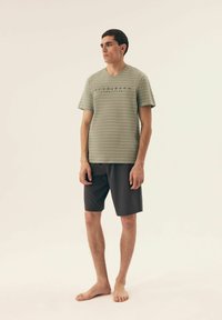 Zielona koszulka w paski z dekoltem w serek i napisem "HENDERSON YACHT CLUB", zestawiona z ciemnymi szortami, model stoi boso na jasnym tle.