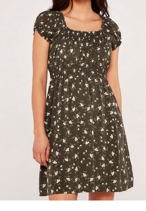 Kobieta ubrana w oliwkowo-zieloną sukienkę z krótkim rękawem, ozdobioną małym białym kwiatowym wzorem, z marszczonym dekoltem i detalami w talii.