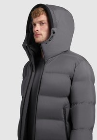 Szara kurtka puchowa z wysokim kołnierzem i kapturem, pikowany wzór, gładka faktura oraz zamek błyskawiczny z przodu. Kieszenie boczne mają ukryte zamki.