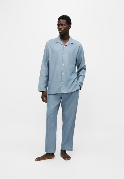 Homme portant une chemise à manches longues boutonnée bleu clair assortie et un pantalon, debout pieds nus devant un fond blanc uni.