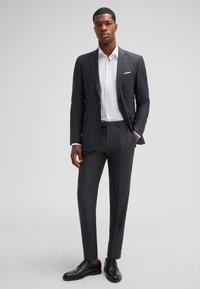Dunkelgrauer Anzug mit schlanker Passform, kombiniert mit einem weißen Hemd. Verfügt über ein Revers mit Kerbe, maßgeschneiderte Hosen und schwarze Lederschuhe.