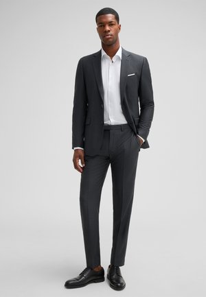 Ciemnoszary garnitur o wąskim kroju, zestawiony z białą koszulą. Garnitur posiada klapę z wycięciem, dopasowane spodnie i czarne skórzane buty.