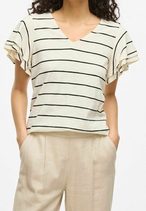 Vrouw draagt een wit shirt met korte mouwen met zwarte horizontale strepen en beige broek met hoge taille, handen in de zakken.