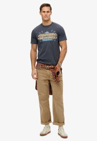 Camiseta gráfica navy con el estampado "Superdry", pantalones cargo beige y una camisa de cuadros atada en la cintura. Zapatillas blancas y beige completan el look.