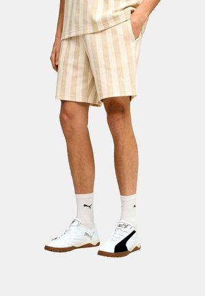 Homme portant un short rayé beige et crème, des chaussettes blanches Puma et des baskets blanches Puma avec des accents noirs, debout avec une main dans la poche.