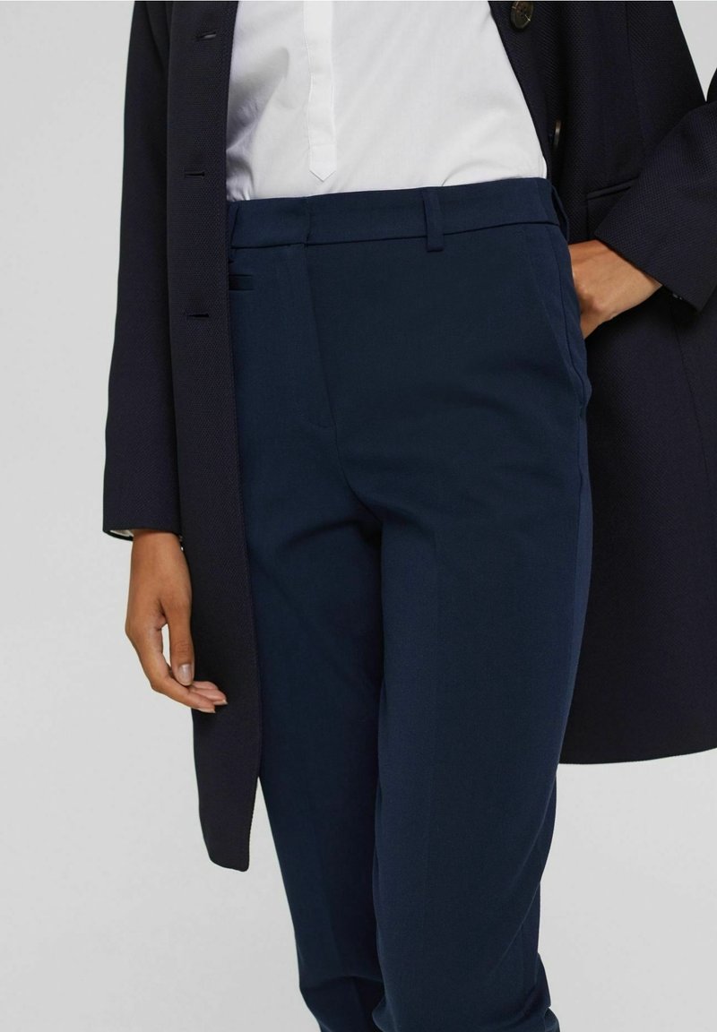 Personne portant un pantalon bleu marine, un chemisier blanc et un manteau bleu marine foncé, une main dans la poche et l'autre détendue sur le côté.