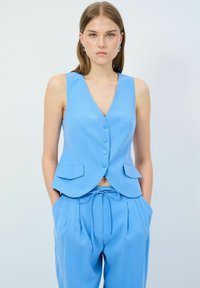 Jasnoniebieska bezrękawnik z głębokim dekoltem w kształcie litery V, pięcioma guzikami i dwoma przednimi kieszeniami, w połączeniu z pasującymi spodniami z pasem na sznurek.